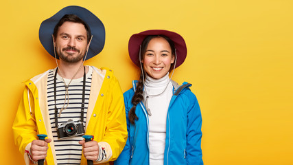 Mixed race trekkers have trip together, stand with trekking poles, carry retro camera, wear hats and casual clothing, have mood for traveling, isolated over yellow background, copy space for promo