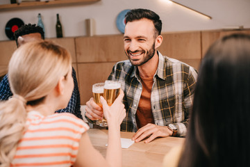 smiling man clinking glasses of light beer with young woman while sitting in pub with multicultural friends