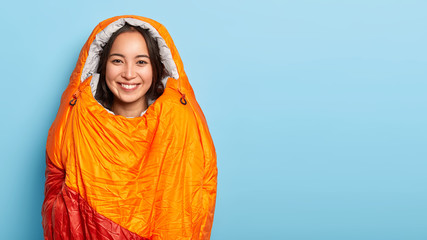 Photo of young Asian girl with toothy smile, stands wrapped in sleeping bag, has adventure trip, spends night in nature, has joyful expression, isolated over blue wall, copy space. Female mountaineer