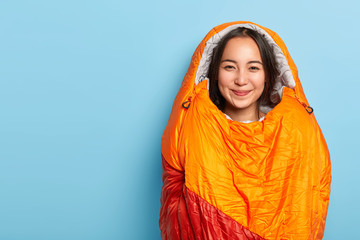 Pleased young Asain brunette woman wrapped in warm orange sleeping bag, spends free time actively, being avtive camper, stands against blue background, blank space for your advertisement or text