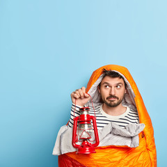 Photo of thoughtful male camper holds gas lamp, stands wrapped in sleeping bag, spends free time on camping, enjoys recreation in calm place at nature, poses against blue studio wall dressed casually