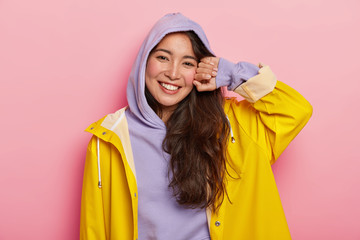 Positive brunette Korean girl in casual hoody and raincoat, smiles pleasantly, being in good mood after outdoor walk with boyfriend, stands against pink background. Cold rainy autumn weather