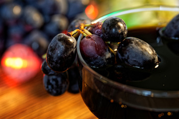 Glass of homemade wine, tincture or juice next to red and black grapes on a background of multi-colored festive lights. Farm harvest on the counter of the fair. Close-up