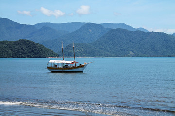 Obraz premium A wooden schooner anchored in a tranquil beach with blue-toned waters and mountains in the background, capturing the serenity and natural beauty of Ubatuba, São Paulo.