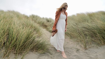young blonde woman looking thoughtful, head down, wearing a long white dress, brown jacket, carrying sandals, descending barefoot sandy sea grass covered hill - Powered by Adobe