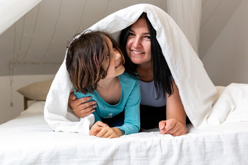 Portrait of mom hugging her daughter under the blanket in the bedroom. Happiness of motherhood concept