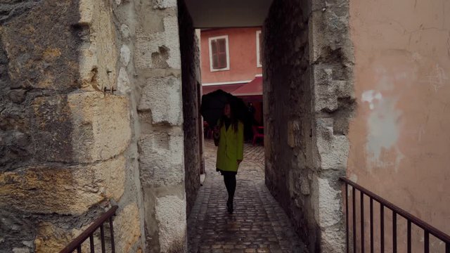 Young brunette girl wearing yellow coat with umbrella walking along narrow cobbled street in Annecy Old Town, France