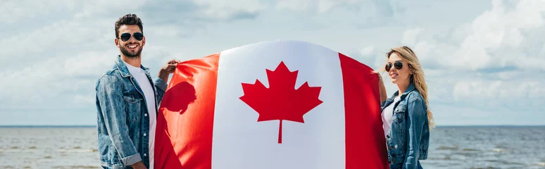 Fotobehang panoramic shot of attractive woman and handsome man smiling and holding canadian flag © LIGHTFIELD STUDIOS