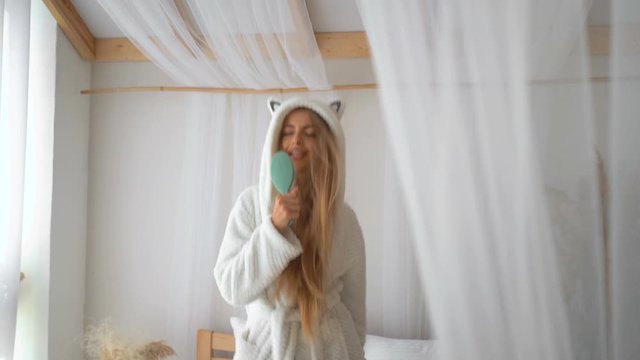 Happy young blonde female with long hair in bathrobe jumping and dancing on bed