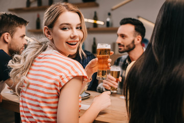 selective focus of cheerful young woman smiling at camera while holding glass of light beer