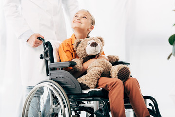 cropped view of pediatrist in white coat and kid with teddy bear on wheelchair