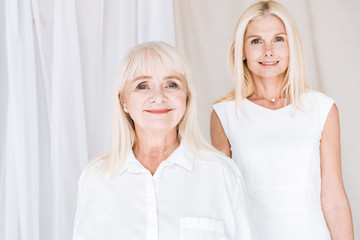 smiling elegant blonde mature daughter and senior mother in total white outfits