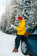 Man in red hat and yellow jacket reading map next to car in forest during winter.