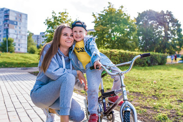 Young happy family smiling happy rejoicing, mom trains child little boy 3-5 years old child, learns first lessons of bicycle, balance, relax, summer autumn day in city park, casual clothes fun.