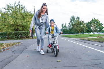 Young mother teaches her son's child, trains balance ride bicycle, little boy 3-5 years old, have rest summer autumn day city park, casual clothes. Caring support help and education, emotions love.