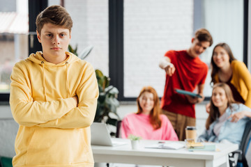 Fototapeta premium sad boy in yellow hoodie with crossed arms and laughing teenagers at desk