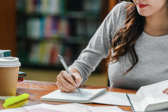 Closeup Asian young Student hand writing homework in library of university or colleage with various book and stationary with coffee cup on wooden table over the book shelf background,Back to school