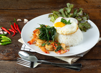 Spicy fried shrimp with basil and chili, serve with rice and egg in white ceramic plate on wood table, Thai style food.