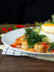 Spicy fried shrimp with basil and chili, serve with rice and egg in white ceramic plate on wood table, Thai style food.