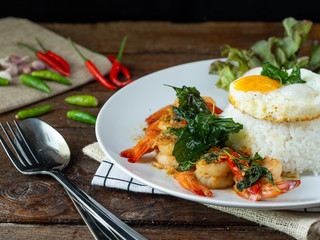 Spicy fried shrimp with basil and chili, serve with rice and egg in white ceramic plate on wood table, Thai style food.
