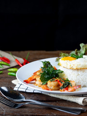 Spicy fried shrimp with basil and chili, serve with rice and egg in white ceramic plate on wood table, Thai style food.