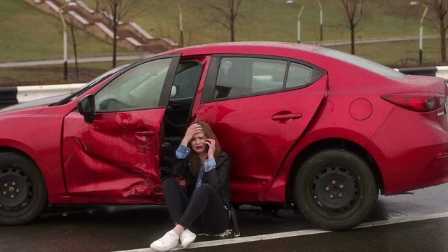 The upset girl sits on the earth near the broken car, other car crashed into her and left. The girl calls on the phone and asks for help, she cries. Car accident in the rain.