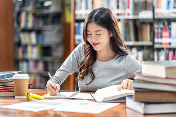 Asian young Student in casual suit reading and doing homework in library of university or colleage with various book and stationary on the wooden table over the book shelf background, Back to school - 292618444