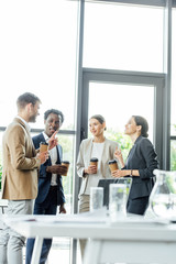 four multiethnic colleagues holding disposable cups of coffee and talking in office