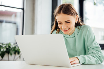 smiling girl in casual hoodie sitting at table and using laptop