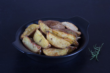 Close up rustic roasted potatoes with rosemary branch on black plate on black background