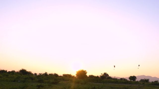 two silhouettes of paramotors are flying from the right to left on clear sky at sunset time