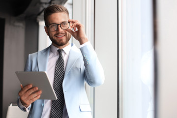 Modern business man in formalwear using digital tablet while standing near window in the office