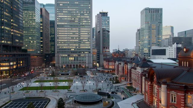 Timelapse zoom out Tokyo railway station old historical buildings and large square with hurrying people among office skyscrapers and with passing by car traffic flow from evening dusk till night