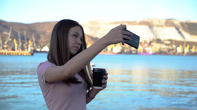Attractive young woman drinking coffee and using mobile and takes selfies on embankment with mountains and cloudy sky on the background