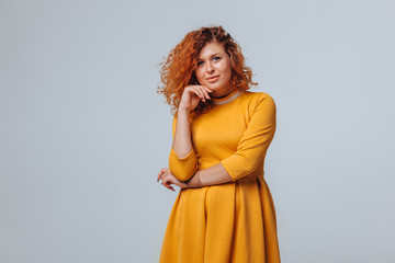 Fototapeta premium A red haired girl in a yellow dress on a light gray background smiles and touches her face