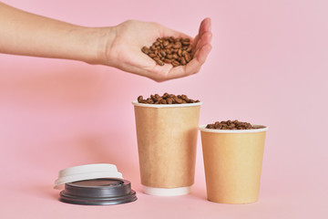 Fototapeta na wymiar Female hand sprinkles coffee beans in coffee paper cup on pink background