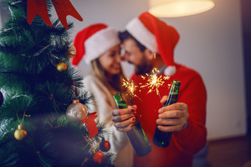 Happy caucasian couple in sweaters and with santa hats on heads cuddling and holding sprinklers and...