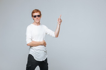 A red-haired man in sunglasses on a light background points. copy space