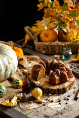 tasty homemade baked pumpkin bundt cake with chocolate on top stands on wooden board on rustic table with assorted colorful small pumpkins and autumn leaves in straw basket