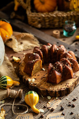 tasty homemade baked pumpkin bundt cake with chocolate on top stands on wooden board on rustic table with assorted colorful small pumpkins and autumn leaves in straw basket