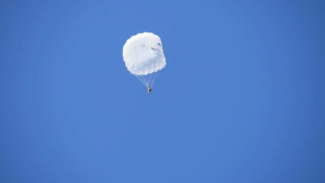 Army man in a white uniform swooping on a white round parachute in a blue sky 