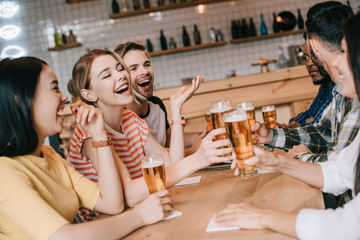 cheerful multicultural friends talking and gesturing while drinking beer in pub together