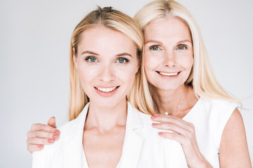 smiling blonde mature mother embracing beautiful young daughter in total white clothes isolated on grey
