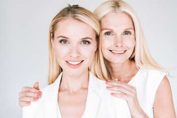 smiling blonde mature mother embracing beautiful young daughter in total white clothes isolated on...