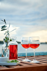 two glasses of rose wine with  olives against blue water on a pier..