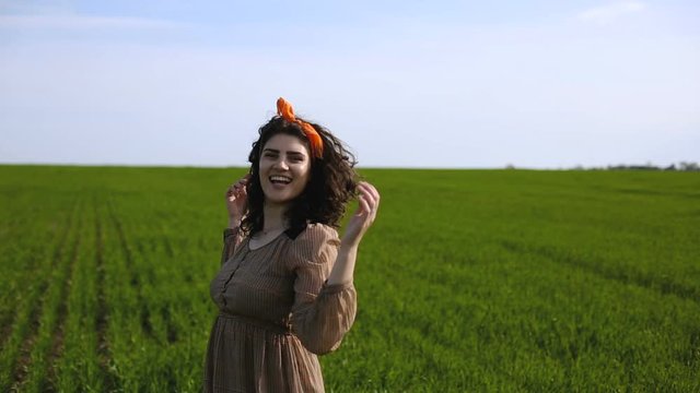 Steady around dolly shot of pretty smiling  young woman with curly hair dressed in brown dress dancing  in the green feeld near the village. Beautiful plus-size model posing.