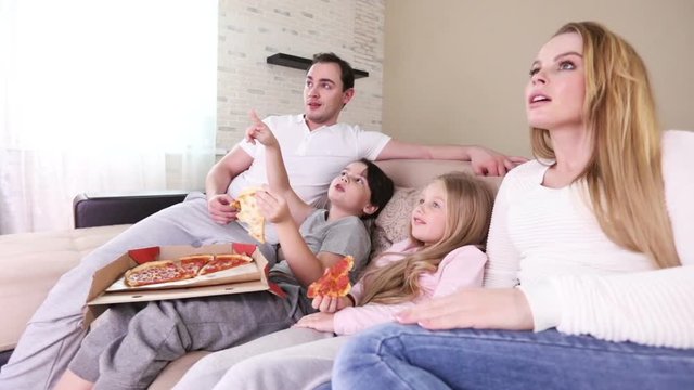 Happy family of parents and two children watching TV on sofa at home and eating pizza