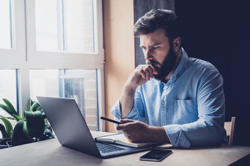 Professional sitting in office in front of laptop. Developer thinking on solutions for work. Home-based student getting distant education. Young serious bearded man in blue shirt working on desktop.