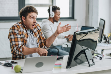 selective focus of shocked programmer gesturing while working near african american colleague