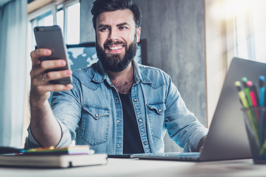 Smiling young man with beard holding smartphone in hand.Digital professional sitting at window in front of laptop.Online education
